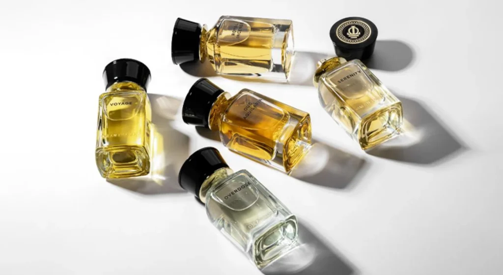 Five popular miniature perfume bottles with black caps, one cap removed, arranged on a white surface. The bottles display different shades of yellow and clear liquid as sunlight casts soft shadows around their elegant shapes.