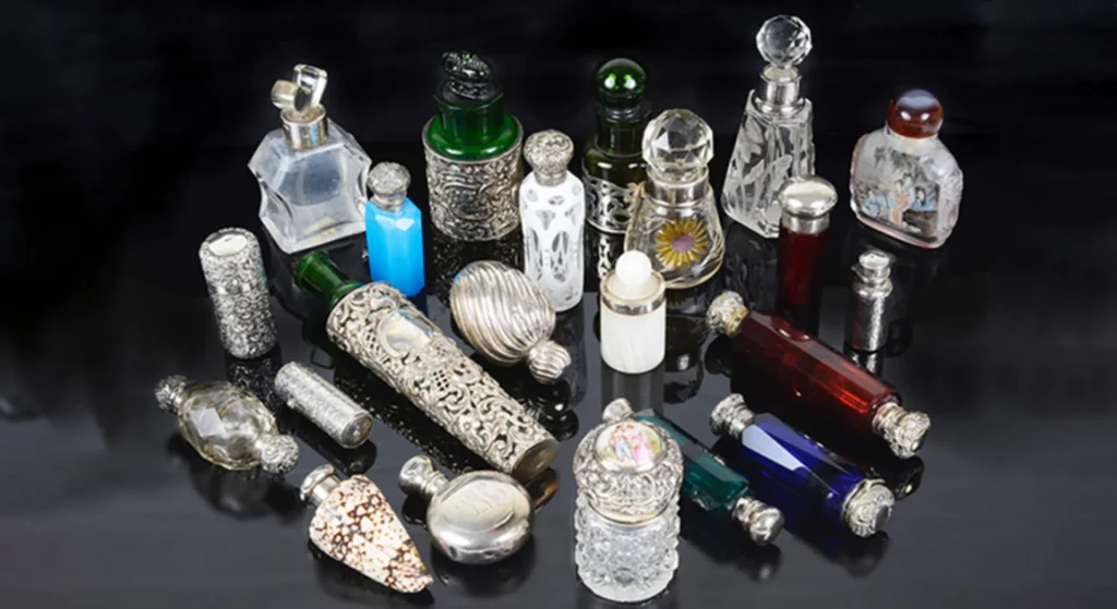 A collection of ornate glass perfume bottles in various shapes, sizes, and colors, some with intricate silver detailing, displayed on a glossy black surface.