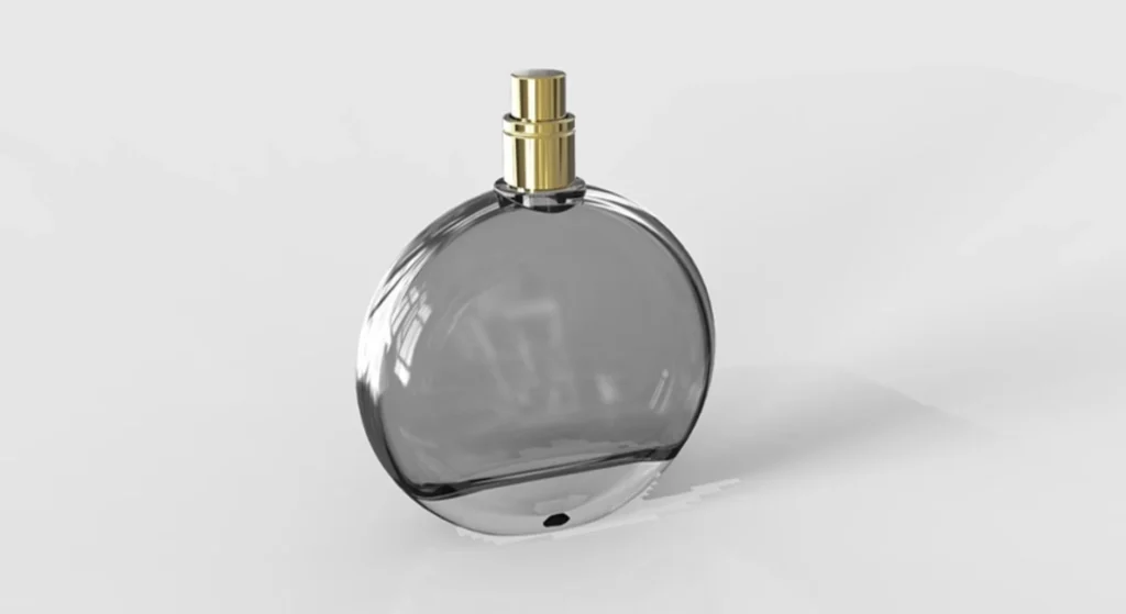 A round, transparent glass perfume bottle with a gold spray nozzle is shown against a plain, light gray background. The empty fragrance vessel reflects light on its surface, highlighting elegant perfume bottle shapes.